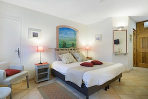 A bedroom with a bed, two bedside tables, a chair, a painting on the wall, and a wooden door. - Capucine Guest Room with Sea View, Wi-Fi, and Air Conditioning (Tourrettes-sur-Loup)