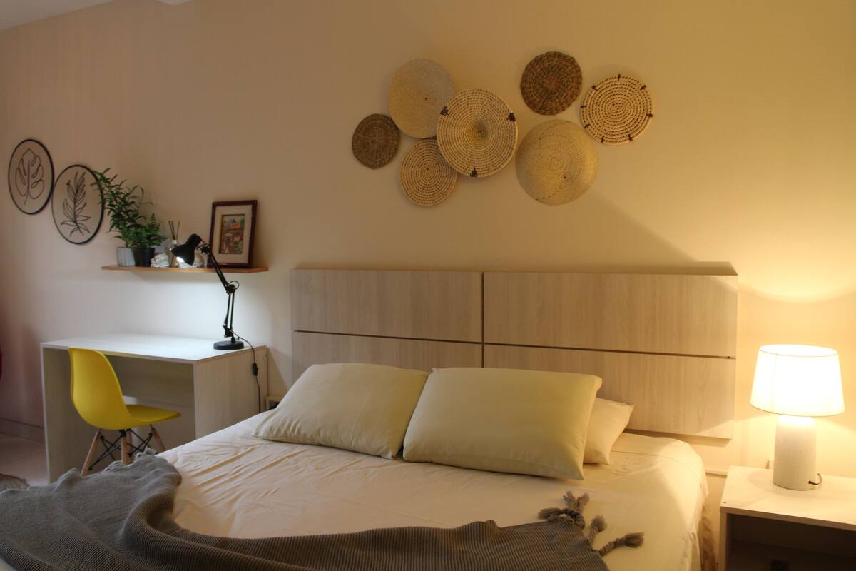 A bedroom with a bed, bedside table, lamp, desk, chair, and wall decorations.
