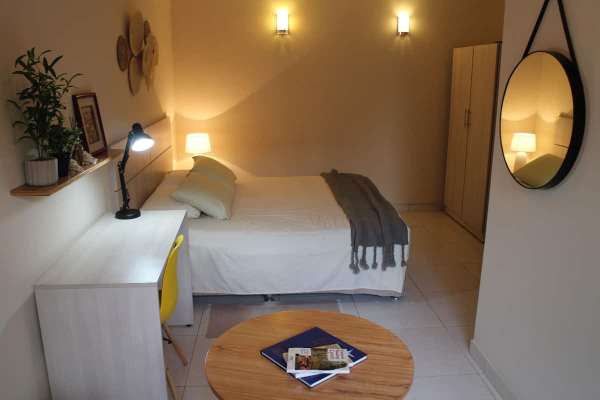 A compact bedroom with a bed, bedside lamps, a desk with a chair, a round wooden table, and a mirror on the wall.