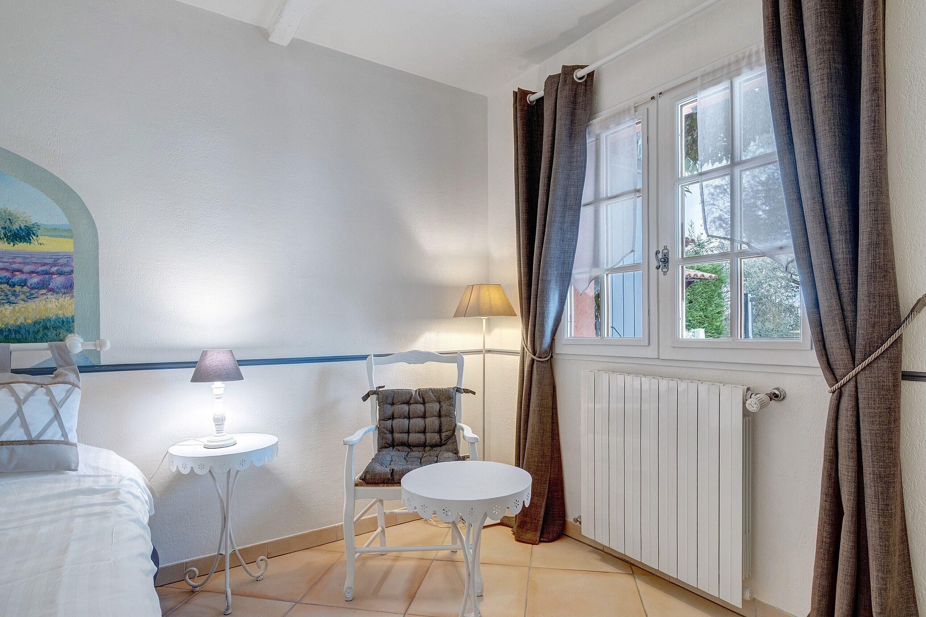 A room with a bed, a chair, a small table, a lamp, a window with curtains, and a radiator.