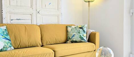 Un canapé jaune agrémenté de coussins à motifs floraux, une table basse en bois avec un livre dessus, un vase en verre abritant des fleurs, un lampadaire et une horloge murale.
