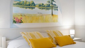 A hotel room with a bed, two yellow pillows, a nightstand with a lamp, and a painting of a landscape on the wall. - Guest Room 'Olive' with Shared Pool, Wi-Fi, and Air Conditioning (Tourrettes-sur-Loup)