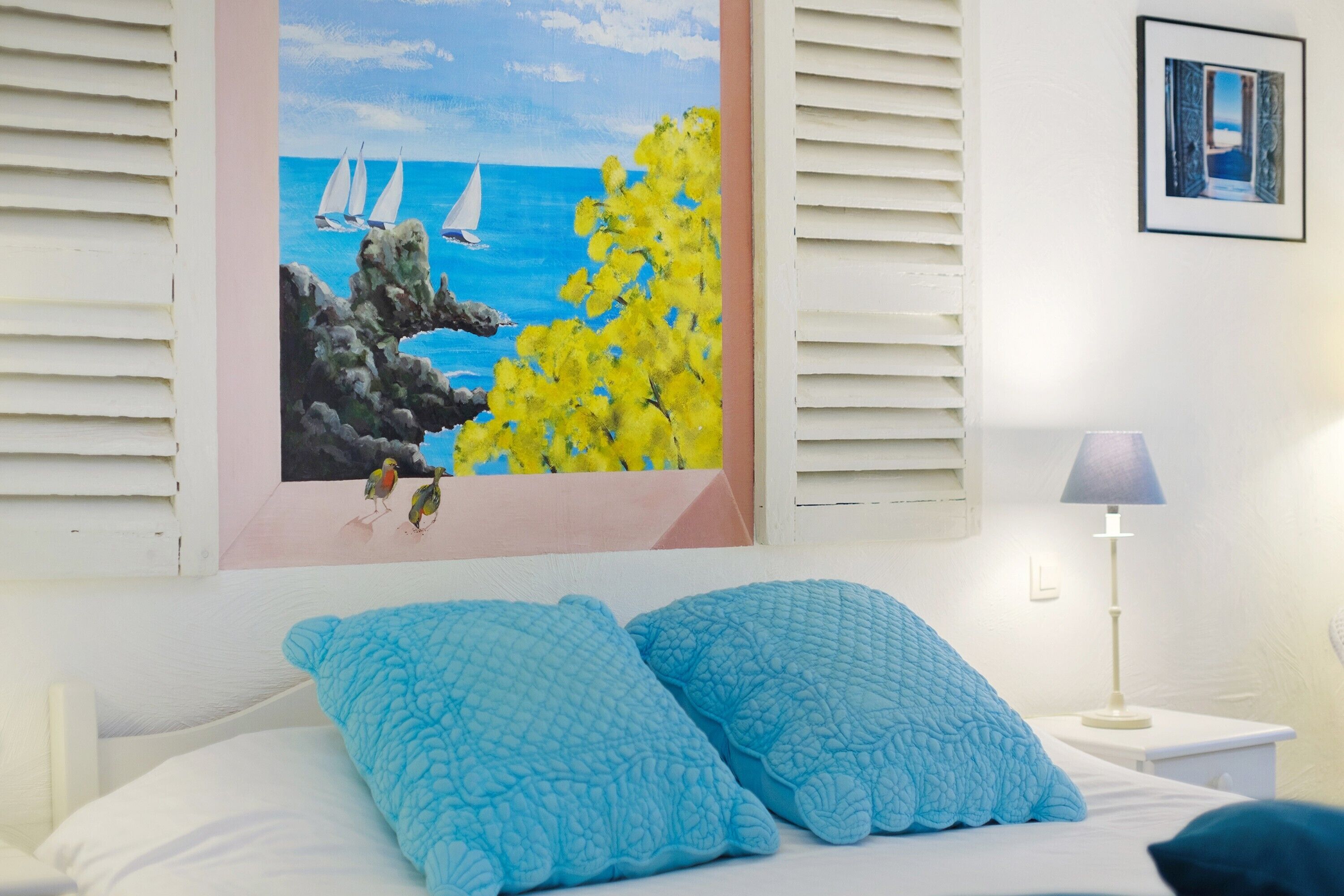 A bedroom with a bed, two blue pillows, a nightstand with a lamp, a painting of sailboats and yellow flowers on the wall, and a framed photo on the wall.