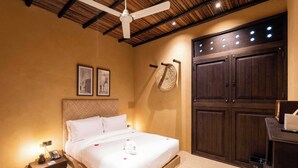 Un lit bien fait, avec une couette blanche et une petite serviette blanche, une table de chevet en bois avec un téléphone et une lampe, et une grande armoire en bois dotée d’un miroir rond.