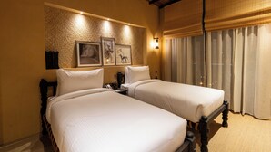 Une chambre d’hôtel avec deux lits, un ventilateur de plafond, une grande fenêtre avec des rideaux et des tableaux encadrés accrochés au mur.