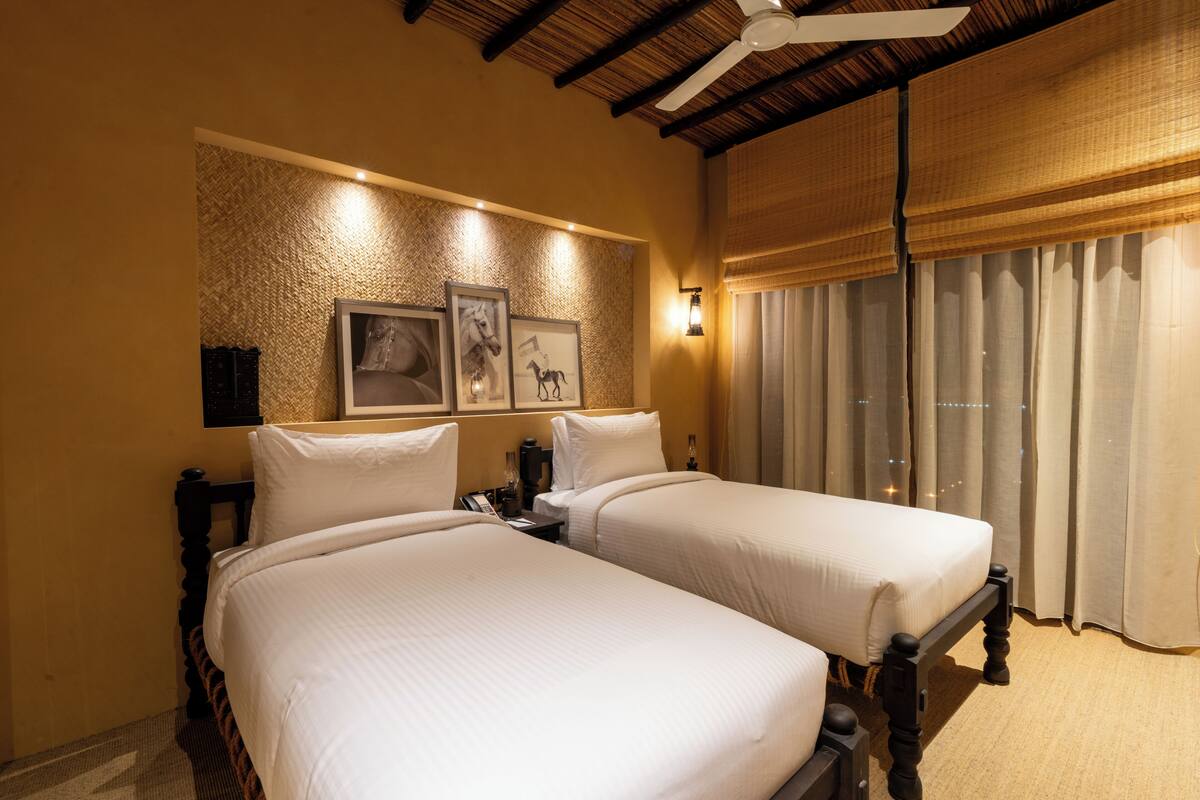 Ein Hotelzimmer mit zwei Betten, einem Deckenventilator, einem großen Fenster mit Vorhängen und gerahmten Bildern an der Wand.