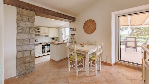 A kitchen with a stone wall, wooden beams, a dining table with chairs, and a view through a glass door.