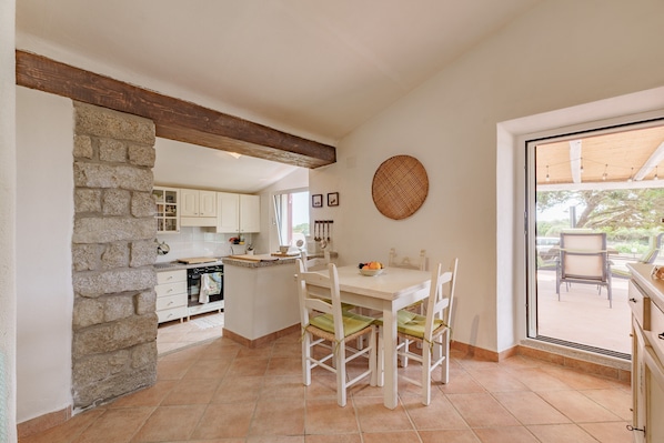 Une cuisine avec un mur en pierre, des poutres apparentes, une table Ă manger avec des chaises et une vue sur lâextĂ©rieur, visible Ă travers une porte vitrĂ©e.
