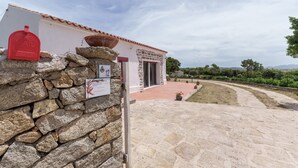 A stone wall with a mailbox and a sign, a building with a red door, and a paved pathway leading to a grassy area.