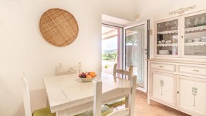 A dining area with a table, chairs, a bowl of fruit, a woven wall hanging, and a cabinet with glass doors.