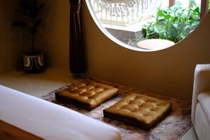 Une pièce comprenant un lit, deux poufs capitonnés, une plante en pot et une vue sur la verdure par une fenêtre.