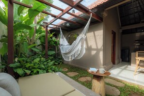 Un hamac suspendu sous une pergola en bois, une petite table ronde avec un livre et une tasse, et un canapé dans un coin du jardin, agrémenté de pierres plates servant de marchepied et d’une végétation luxuriante.