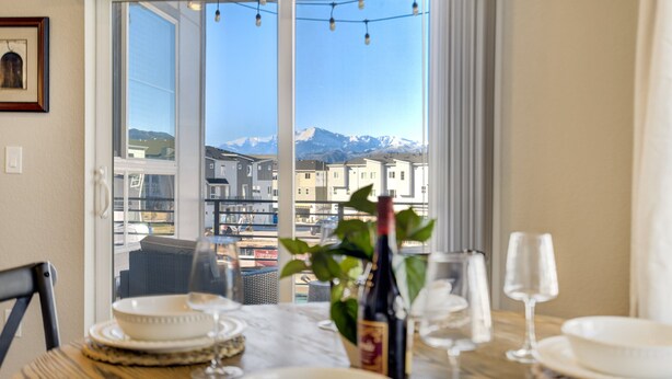 Una mesa de comedor con platos, vasos y una botella, frente a una ventana con vistas a edificios y montañas.