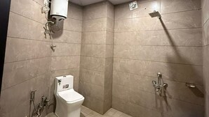A small, tiled bathroom with a toilet, a shower, and a blue bucket. - SM Hotel (Ghaziabad)