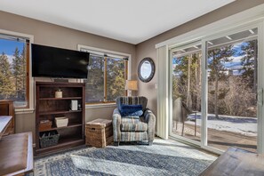A living room with a large flat-screen TV, a bookshelf, a patterned armchair, and a view of a snowy outdoor deck. - Cozy 1BR Near Lifts + Hot Tub Access (Breckenridge)