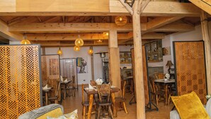 Um interior rústico com vigas de madeira aparentes, área de jantar com cadeiras e mesas de madeira, e luminárias pendentes.