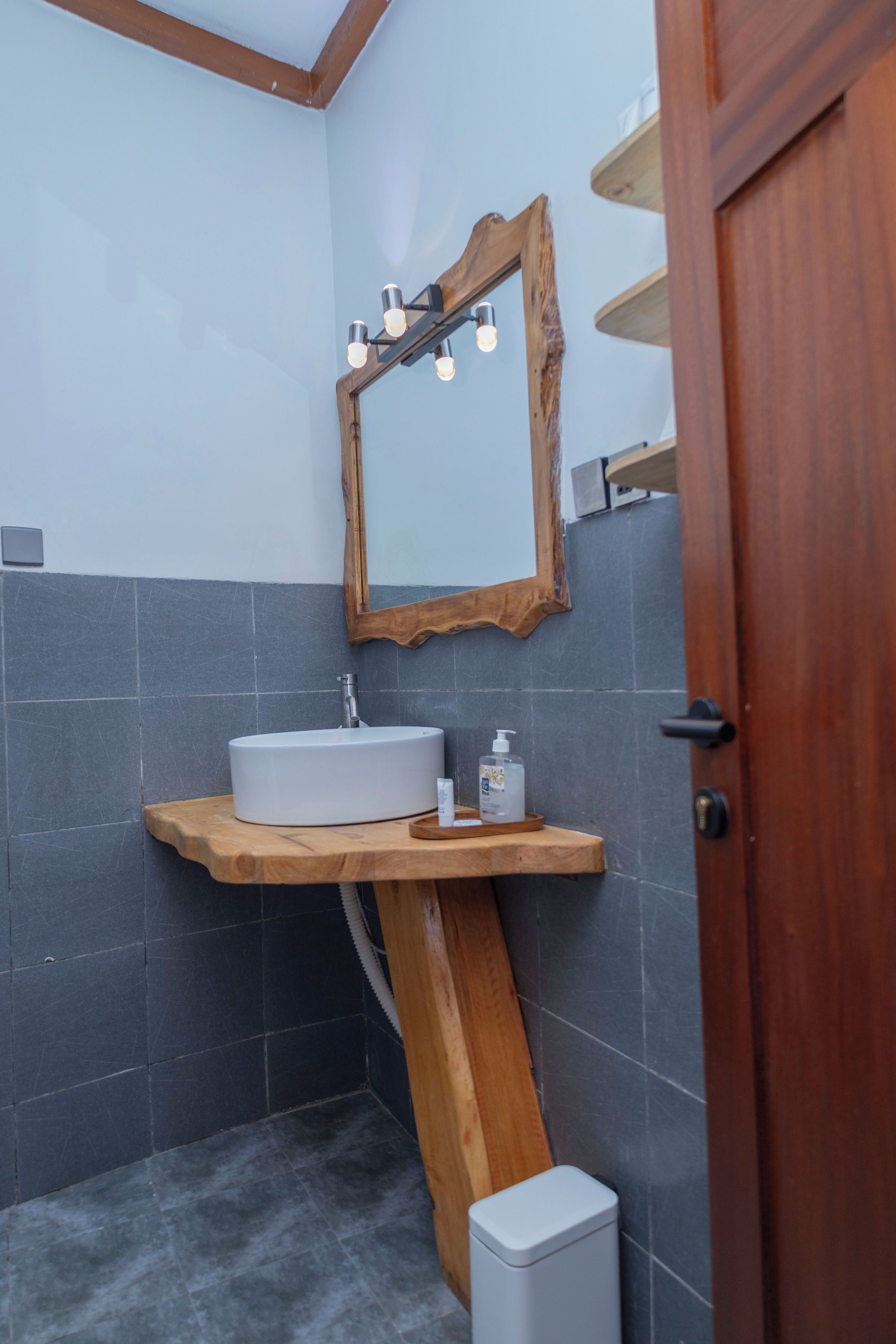 A bathroom with a wooden vanity, a white sink, a mirror with lights, and a door.