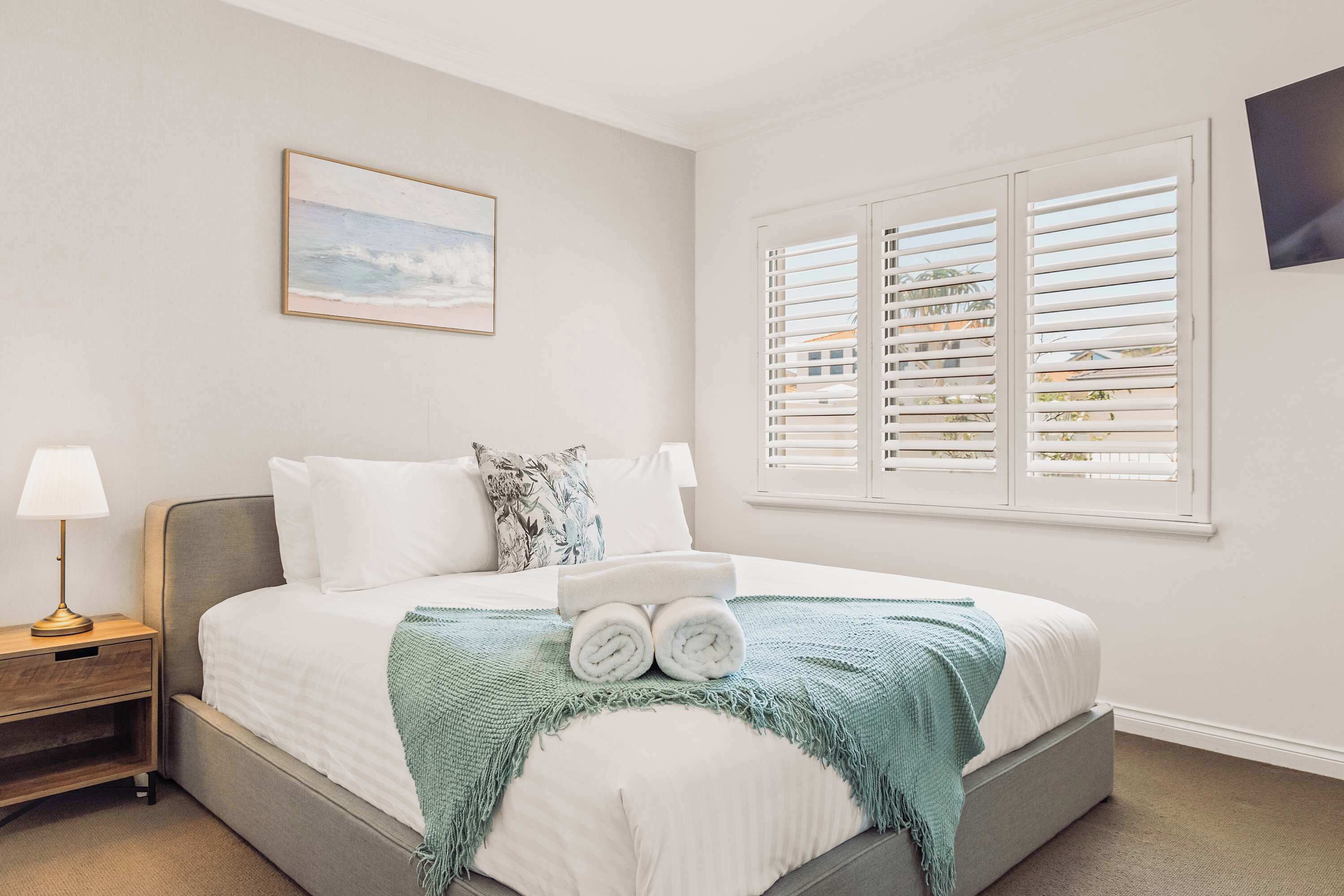A neatly made bed with rolled towels, a bedside table with a lamp, and a window with blinds.
