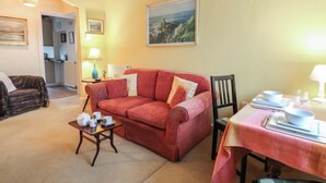 A cozy living room with a red sofa, a small wooden coffee table, and a dining table with a tablecloth. There is a painting on the wall and a lamp on a side table. - 6 Church Lane (Llanidloes)