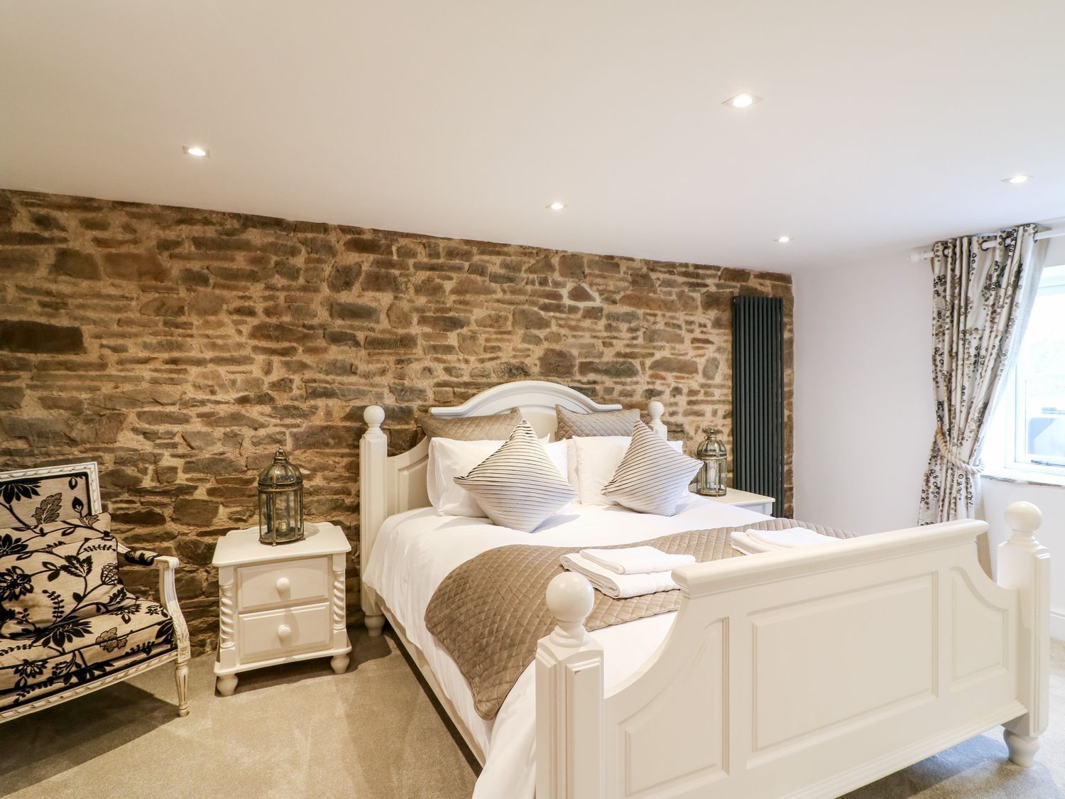 A bedroom with a stone wall, a white bed, and a nightstand with a lamp.