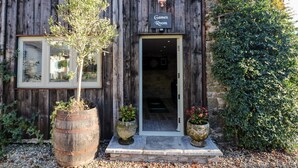 A rustic building with a wooden facade, a sign reading "Games Room," and a door leading inside. There are two potted plants on the steps and a large barrel planter to the left. - Lake View (Crediton)