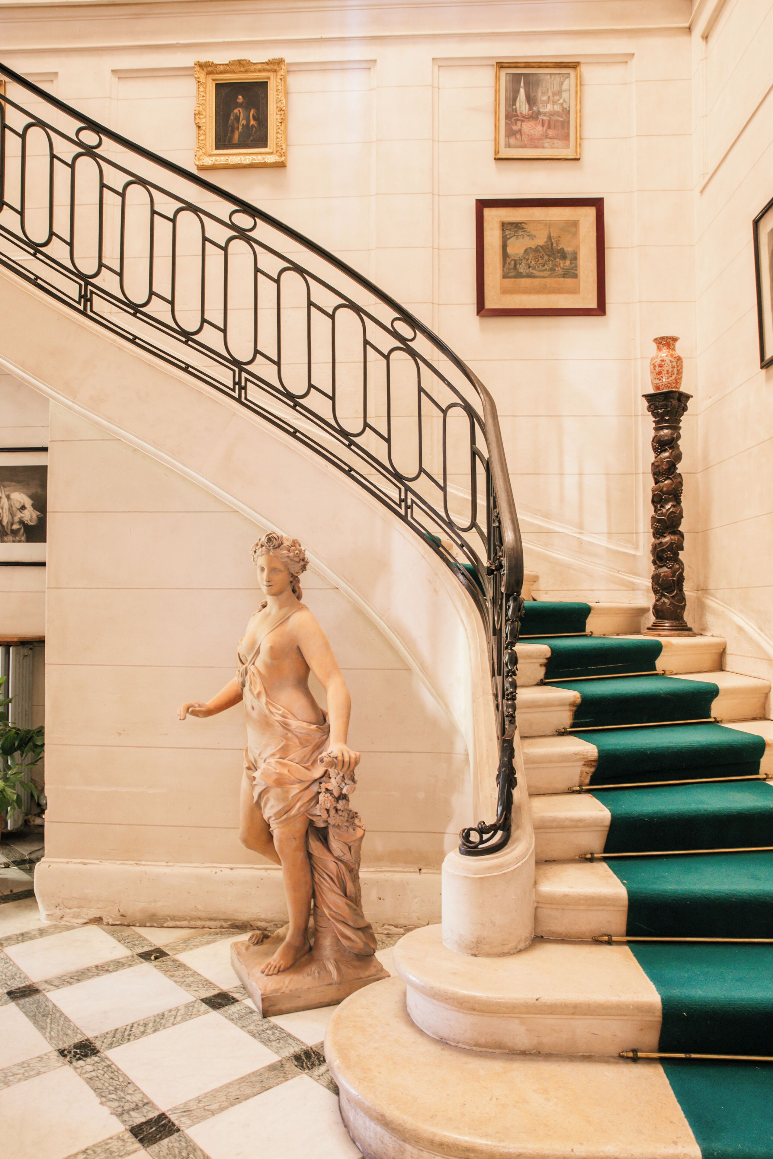 A marble staircase with green carpeted steps, a statue at the base, and framed artwork on the walls.