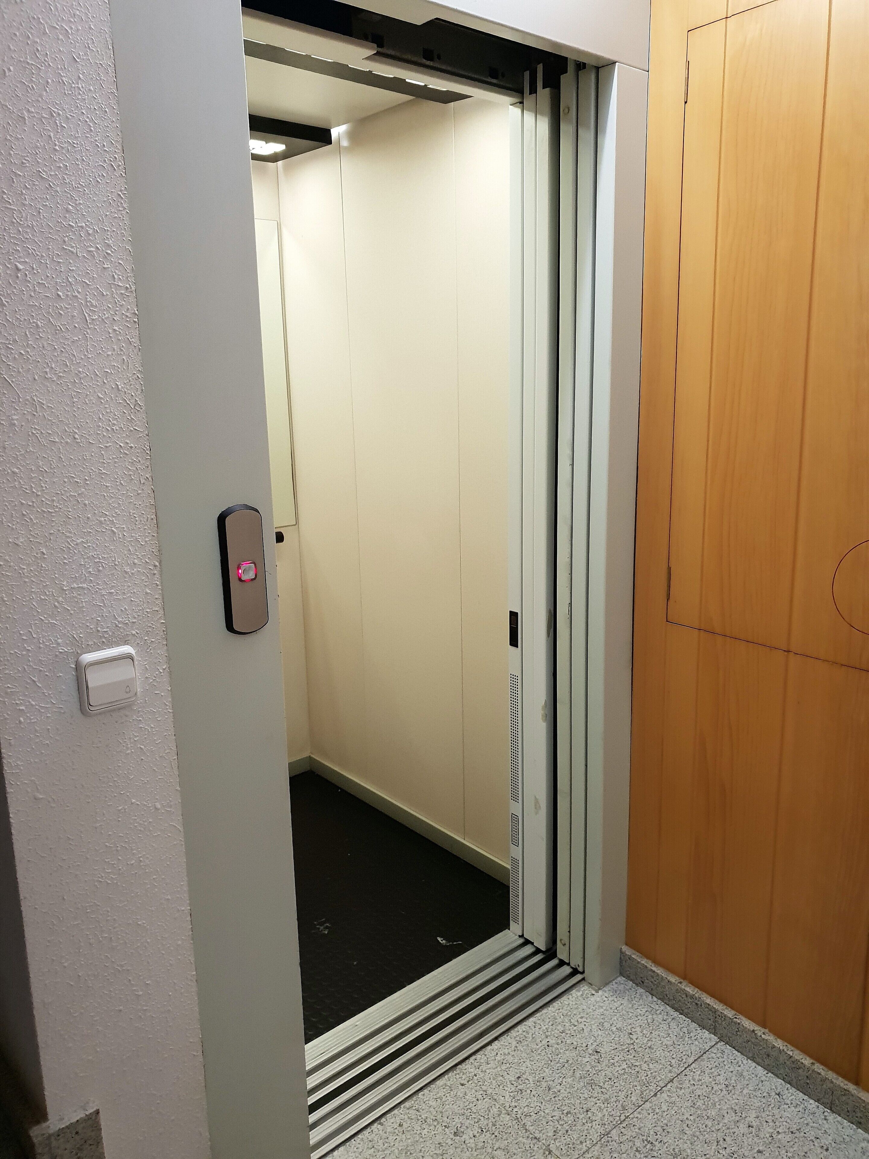 An elevator with a metallic door, a control panel with a red button, and a wooden door adjacent to it.