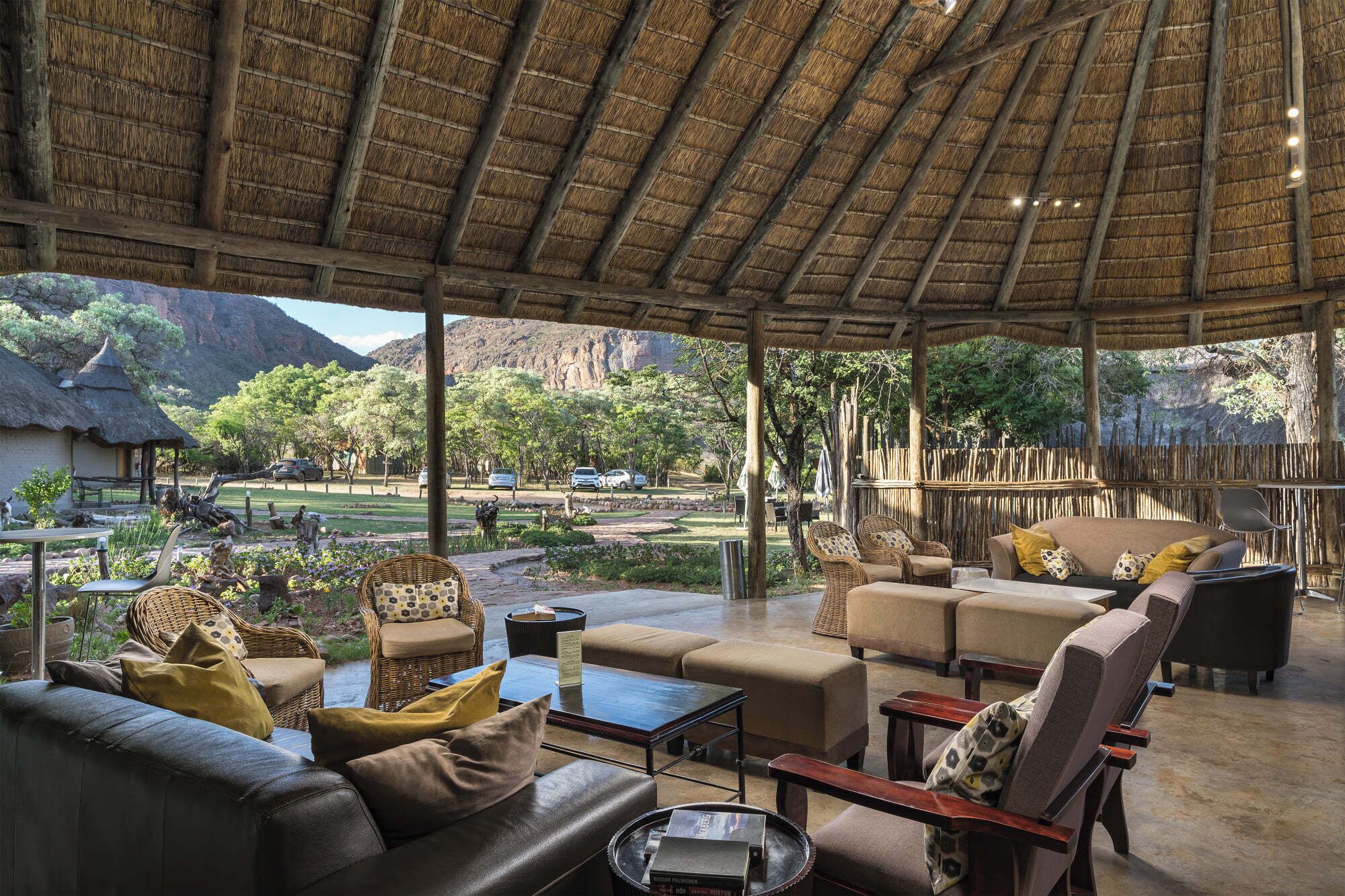 An open-air lounge with a thatched roof, furnished with sofas and chairs, overlooking a landscaped garden and a mountainous backdrop.