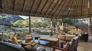 An open-air lounge with a thatched roof, furnished with sofas and chairs, overlooking a landscaped garden and a mountainous backdrop.