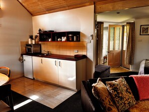 A compact kitchen with a refrigerator, oven, and microwave, a dining area with a chair, and a living space with a sofa and a patterned cushion.