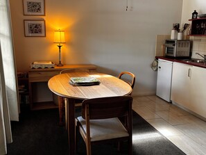 A dining area with a wooden table and chairs, a lamp, a microwave, and a refrigerator in the background.