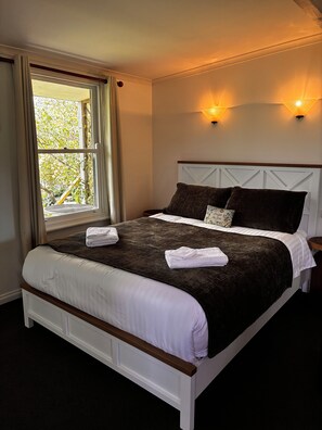 A neatly made bed with white and brown linens, a window with curtains, and wall-mounted lights.
