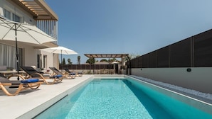 A swimming pool with lounge chairs and umbrellas, surrounded by a modern building with a covered patio area. - Villa Luna d'oro (Zakynthos)