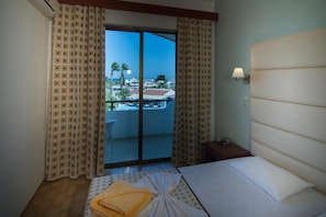 Ein Hotelzimmer mit einem Bett, einem Nachttisch, einer Lampe und Blick auf einen Balkon mit Pool und Gebäuden.