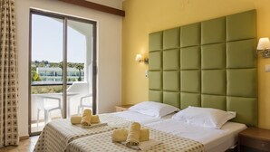 A hotel room with two beds, a green upholstered headboard, a balcony with white chairs, and a view of greenery. - Apartment in A Hotel 'Double Room No 2' with Shared Pool, Wi-Fi and Air Conditioning (Faliraki)