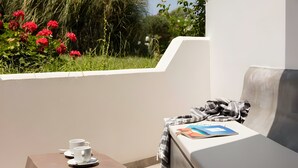 Ein Balkon mit weißer Wand, einem kleinen Tisch mit zwei weißen Tassen, einer Couch mit Decke und Buch sowie roten Blumen im Hintergrund.