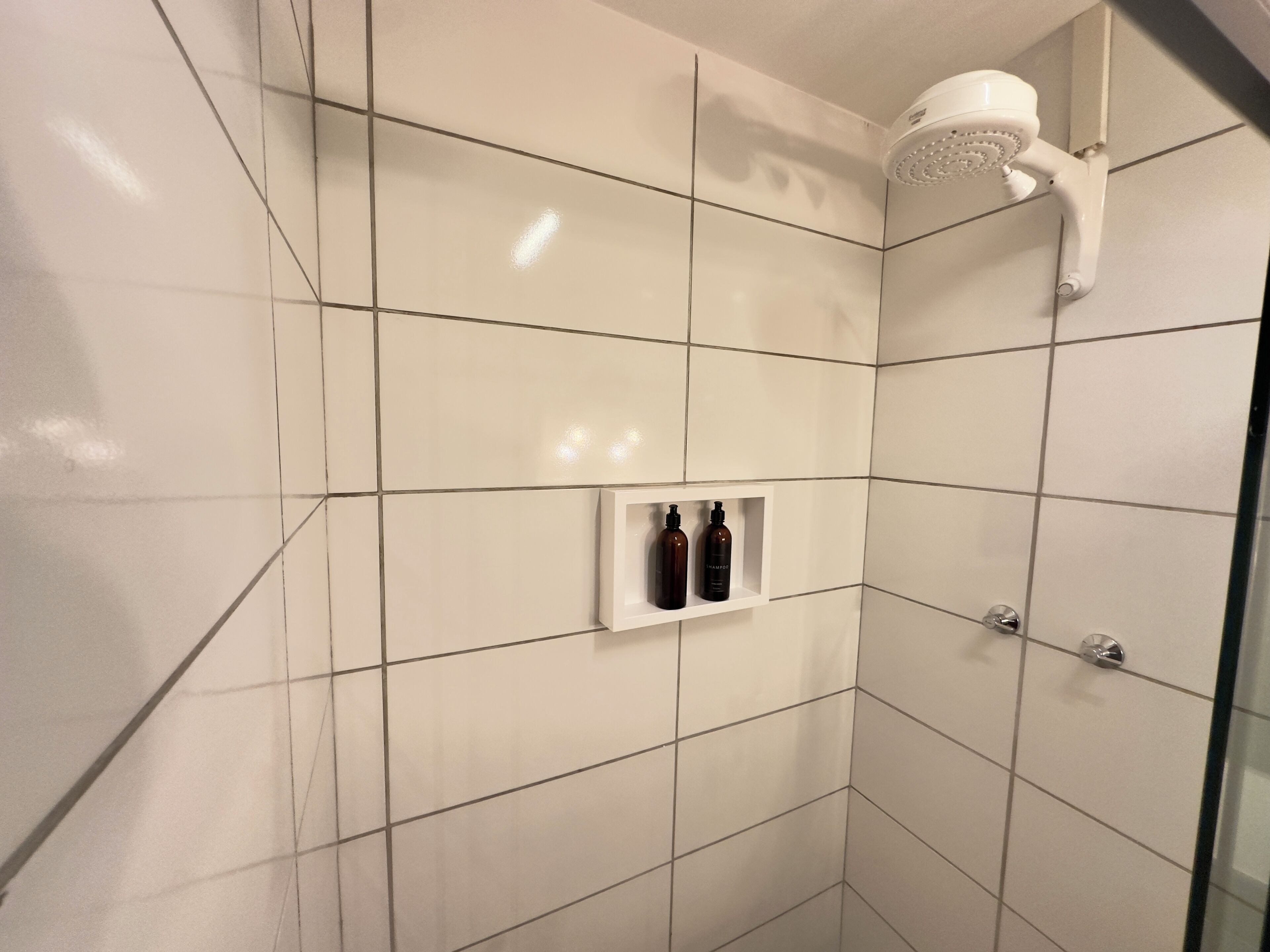 Une salle de bain moderne avec des carreaux blancs, une douche équipée d’un pommeau de douche à main et une étagère sur laquelle sont posés deux flacons de shampoing.