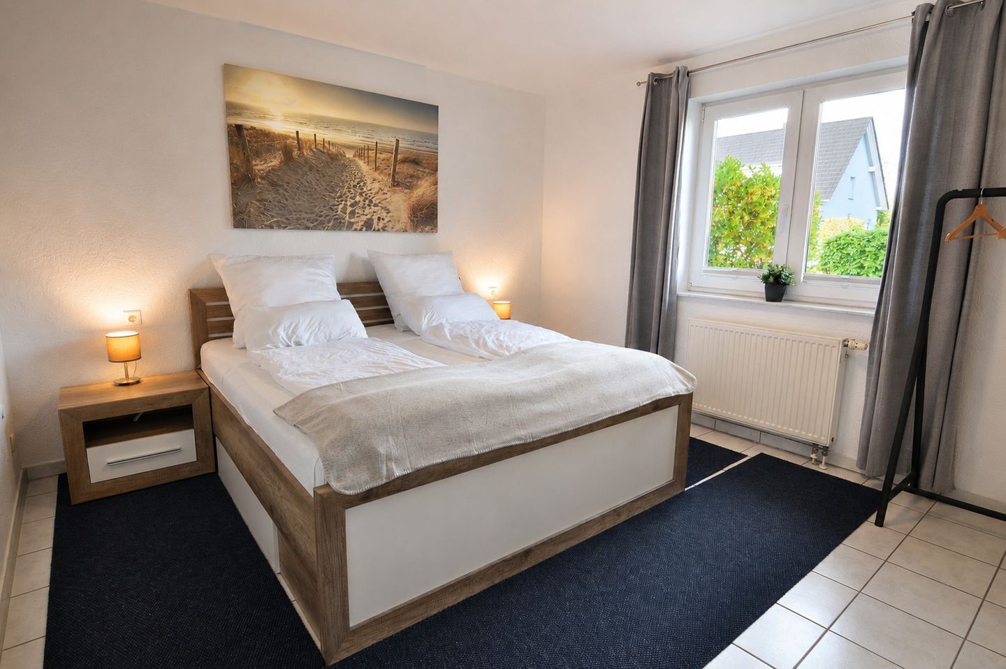 Een slaapkamer met een bed, nachtkastjes, een raam met gordijnen, een radiator en een aan de muur hangend schilderij.