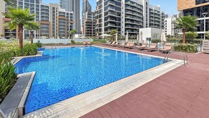 Una piscina con sillones de descanso y palmeras, rodeada de modernos edificios altos.