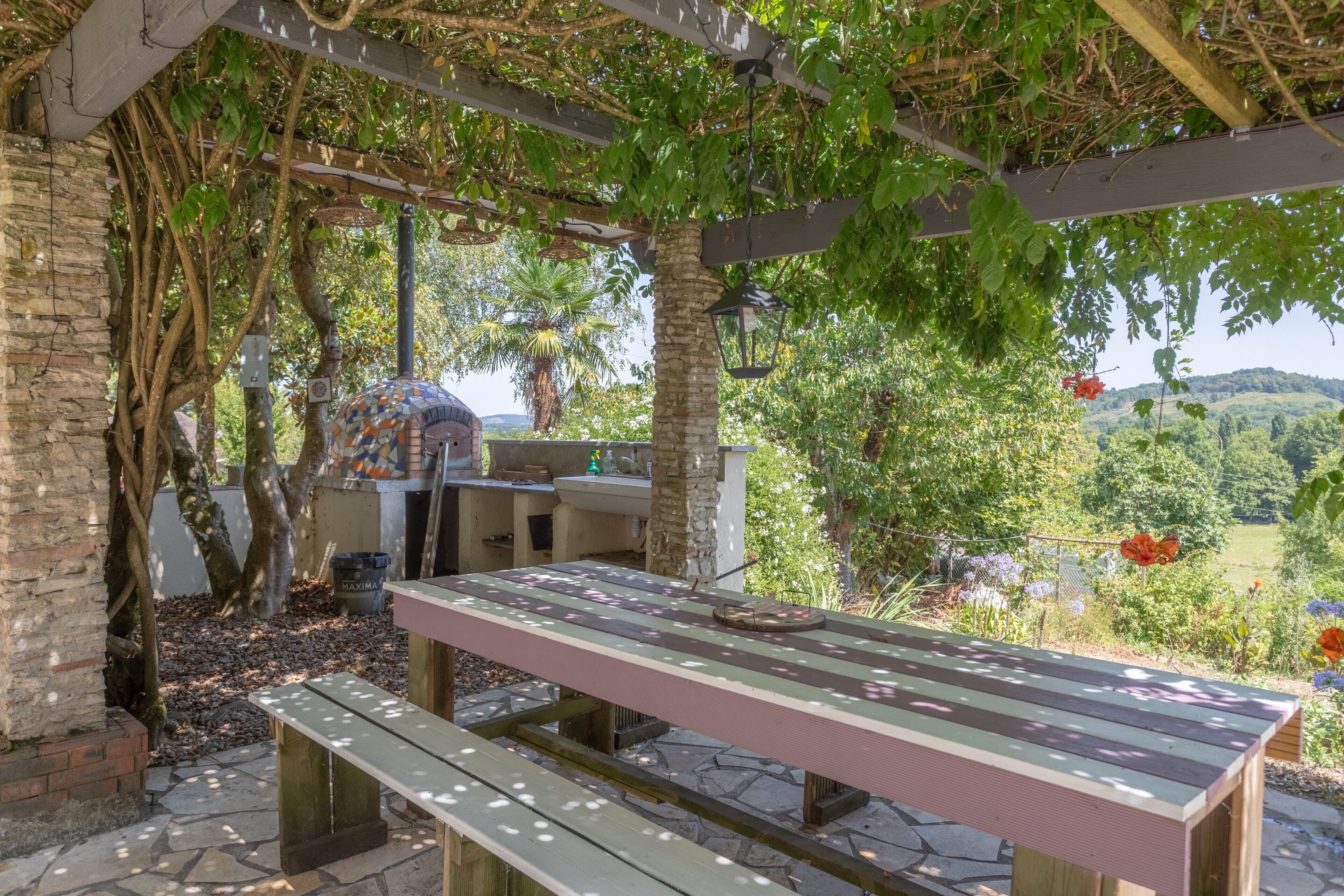 Un espace de restauration extérieur abrité, avec une table et des bancs, entouré d’arbres et offrant une vue sur un paysage vallonné.