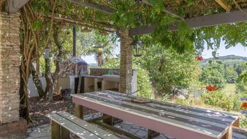 Un espace de restauration extérieur abrité, avec une table et des bancs, entouré d’arbres et offrant une vue sur un paysage vallonné.