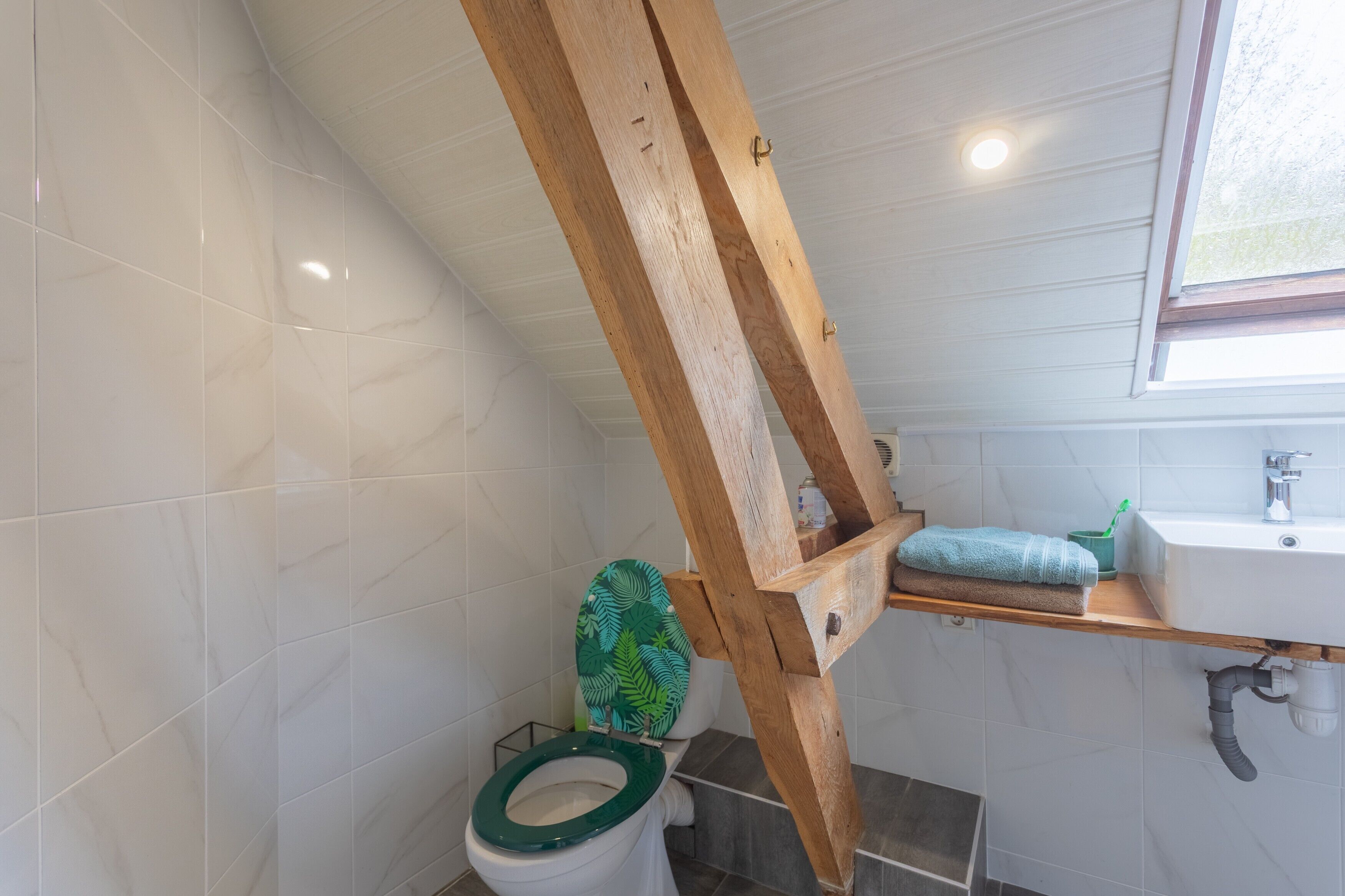Une salle de bain avec une structure en poutres apparentes, des toilettes avec un abattant vert, un lavabo avec un gobelet vert et une fenêtre.