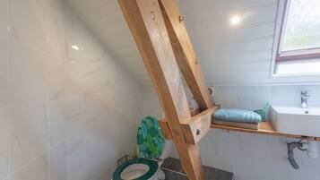 Une salle de bain avec une structure en poutres apparentes, des toilettes avec un abattant vert, un lavabo avec un gobelet vert et une fenêtre.