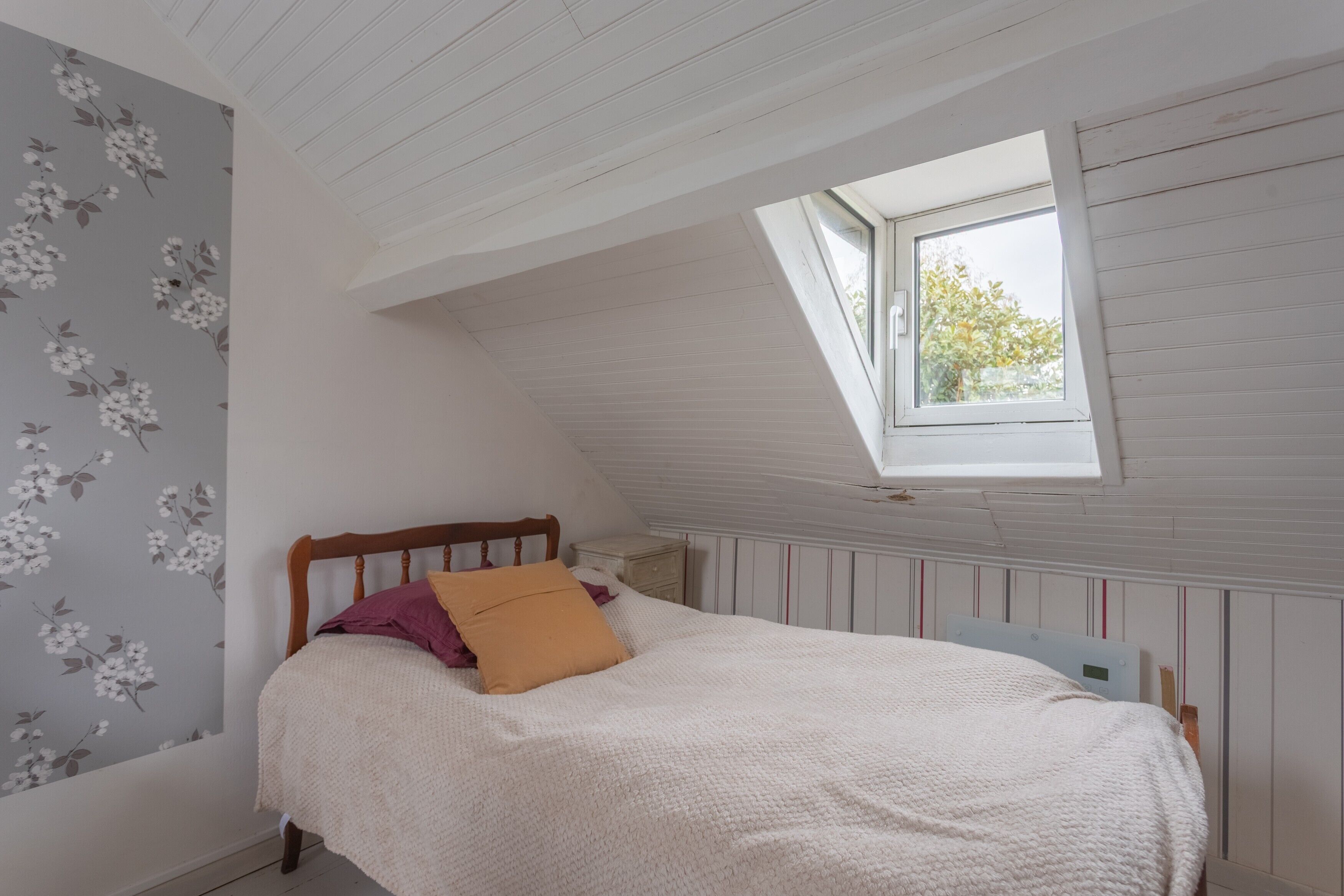 Une chambre avec un plafond en pente, une fenêtre, un lit avec des oreillers, une table de chevet et un papier peint à motifs floraux.