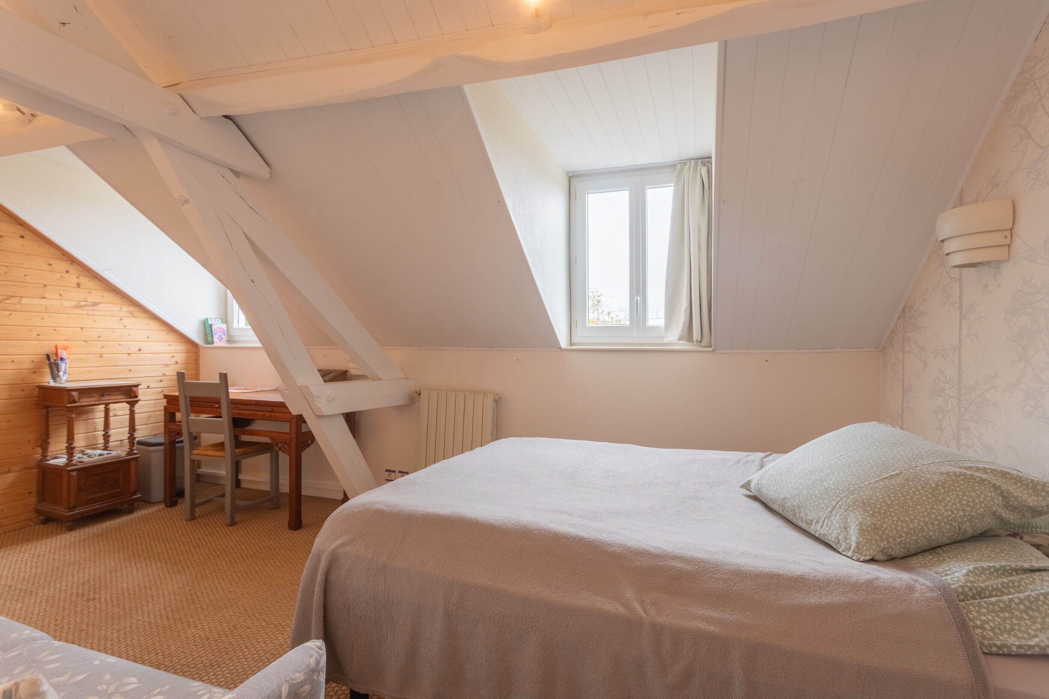 A bedroom with a sloped ceiling, a window, a bed, a chair, and a radiator.
