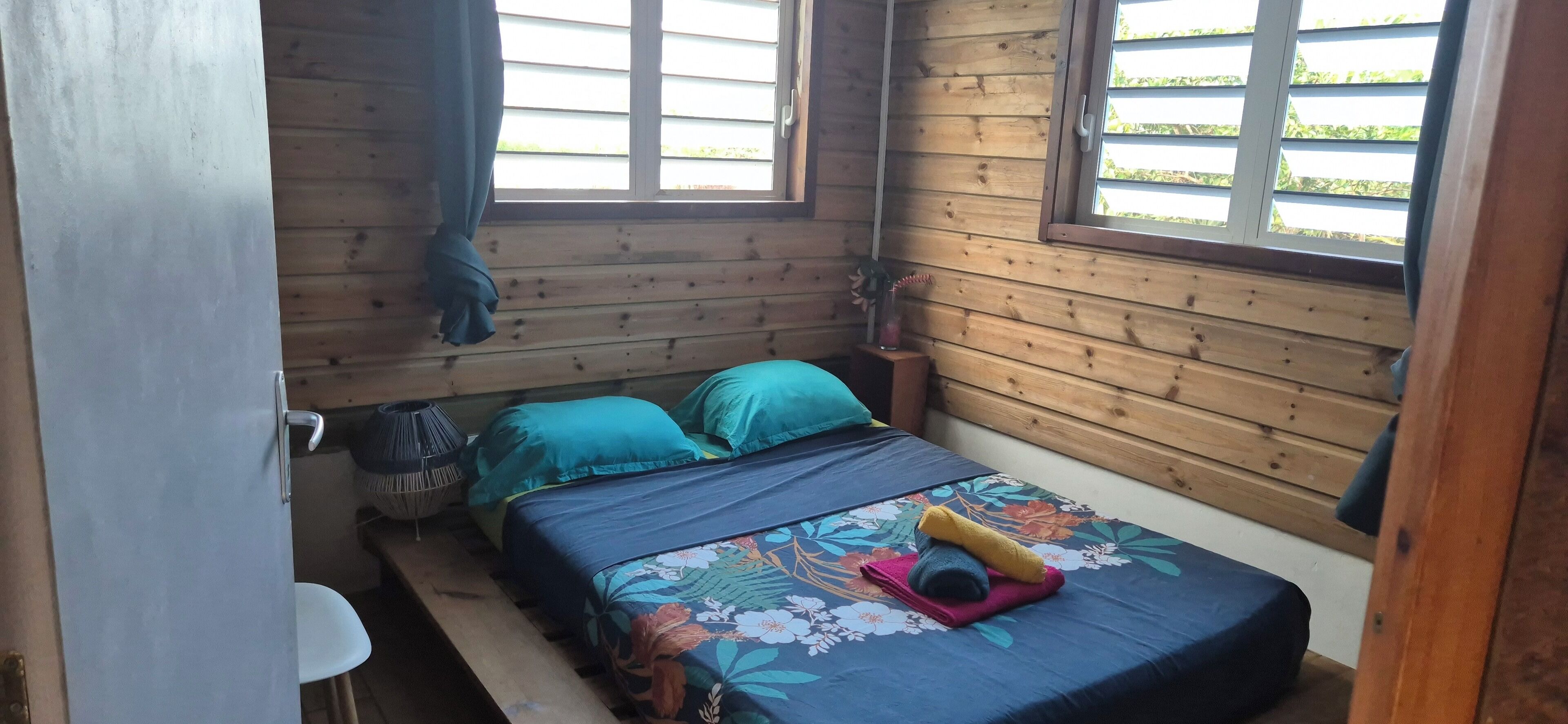 A small, cozy room with a wooden interior, a bed with a floral bedspread, a small table with a chair, and a window with curtains.