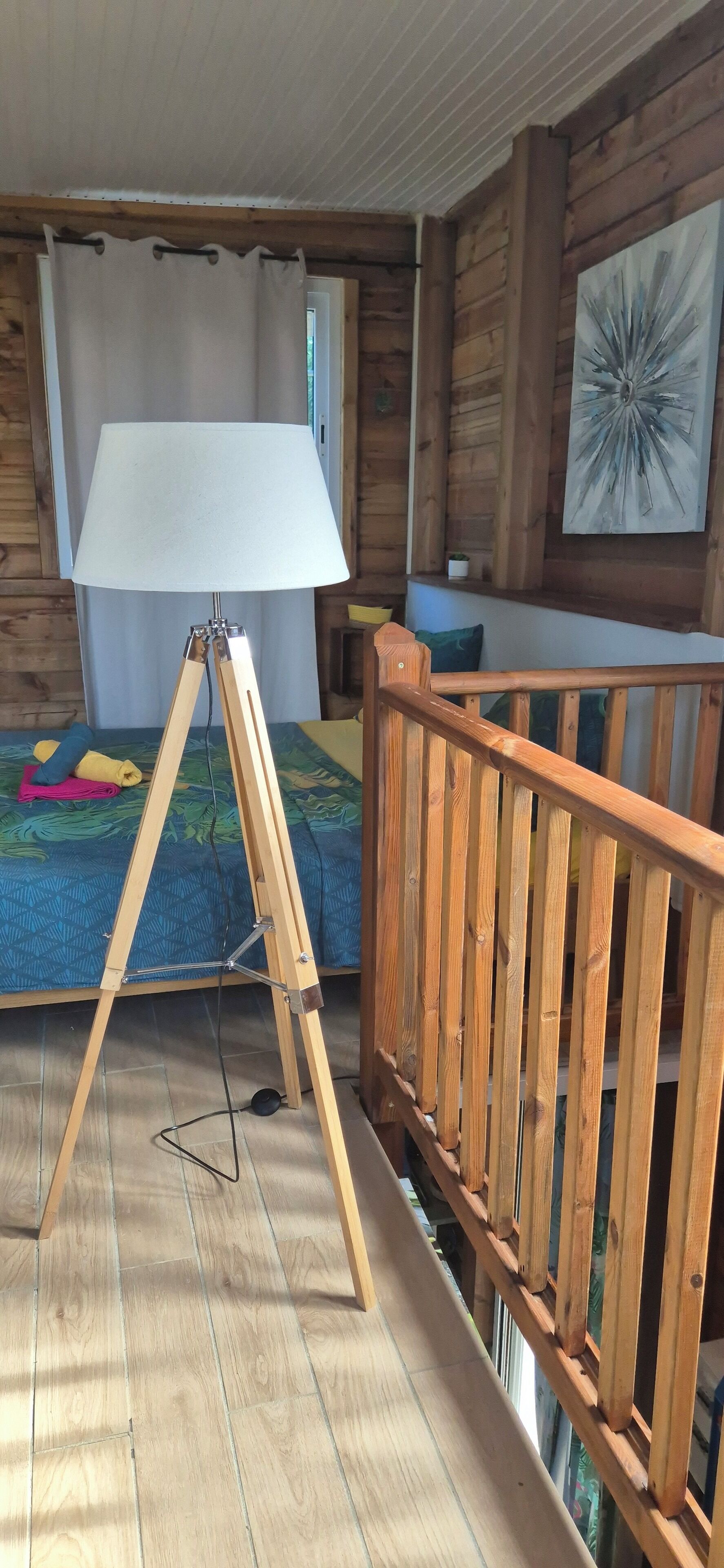 Een kamer met houten vloer, een bed met een blauw bedsprei met patroon, een staande lamp en een houten leuning.