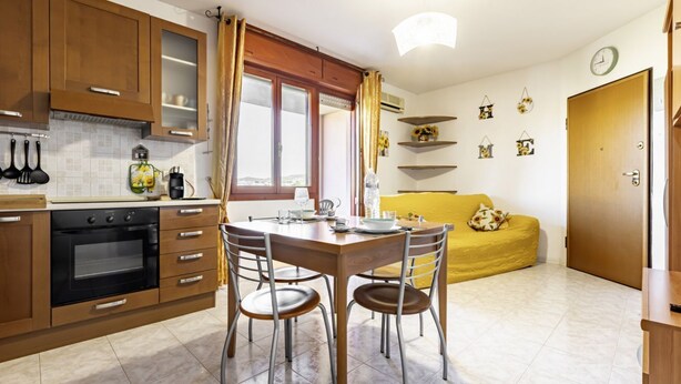 A kitchen with wooden cabinets, a dining table with chairs, a yellow sofa, and a clock on the wall. - GuestHost - Charming apartment of about 65 sqm, perfect for up to 4 guests, located on the third floor of a building with an elevator (NOT for disabled people.)The property is located in Quartu Sant’Elena, in a residential area well served by main amen (Quartu Sant'Elena)