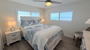 Un dormitorio con cama, mesitas de noche, ventilador de techo y dos ventanas con persianas.
