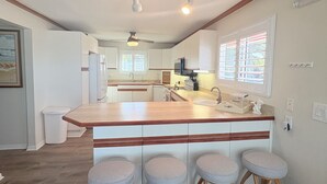 Una cocina con una isla central, taburetes de bar, un ventilador de techo y una ventana con persianas.